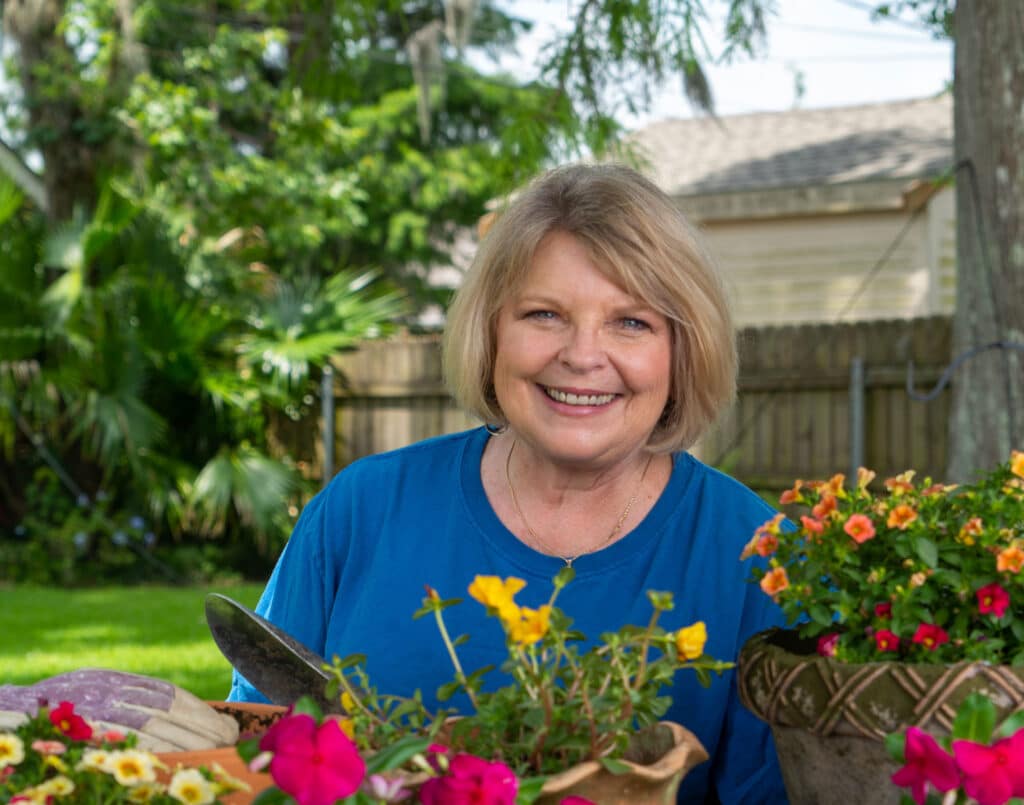 photo-1 A woman in a blue shirt standing in front of a pot of flowers.