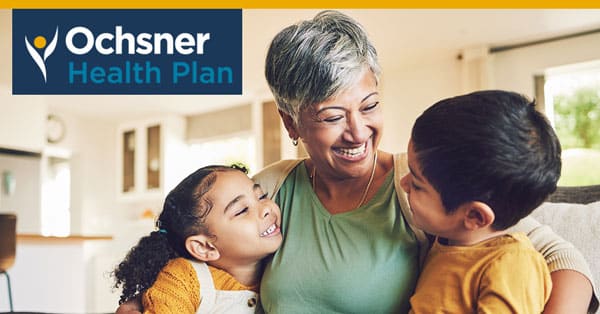 An Older Woman Is Sitting On A Couch, Smiling, And Hugging A Young Girl And Boy. The Ochsner Health Plan Logo Is Displayed In The Upper Left Corner.