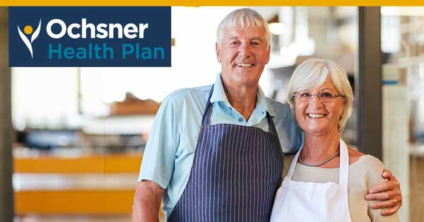 Two smiling elderly individuals wearing aprons stand together. A sign that reads “Ochsner Health Plan” is located in the upper left corner of the image. Two Smiling Elderly Individuals Wearing Aprons Stand Together. A Sign That Reads "ochsner Health Plan" Is Located In The Upper Left Corner Of The Image.
