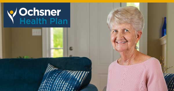 An Elderly Woman In A Pink Sweater Sits On A Couch With Blue And White Pillows. The Ochsner Health Plan Logo Is Displayed In The Upper Left Corner.