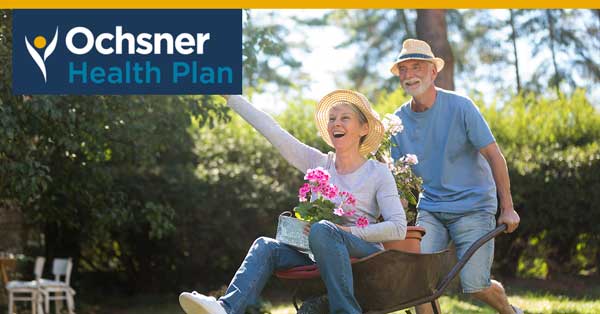 A Man Pushes A Woman Sitting In A Wheelbarrow With Flowers; Both Are Wearing Hats And Smiling. "ochsner Health Plan" Logo Is In The Top Left Corner.