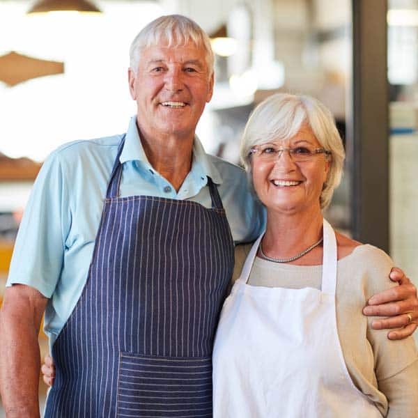 A senior man and woman, both wearing aprons, smile and pose together indoors, showcasing the joy they find in spending time together while discussing Our Plans for their future. A Senior Man And Woman, Both Wearing Aprons, Smile And Pose Together Indoors, Showcasing The Joy They Find In Spending Time Together While Discussing Our Plans For Their Future.