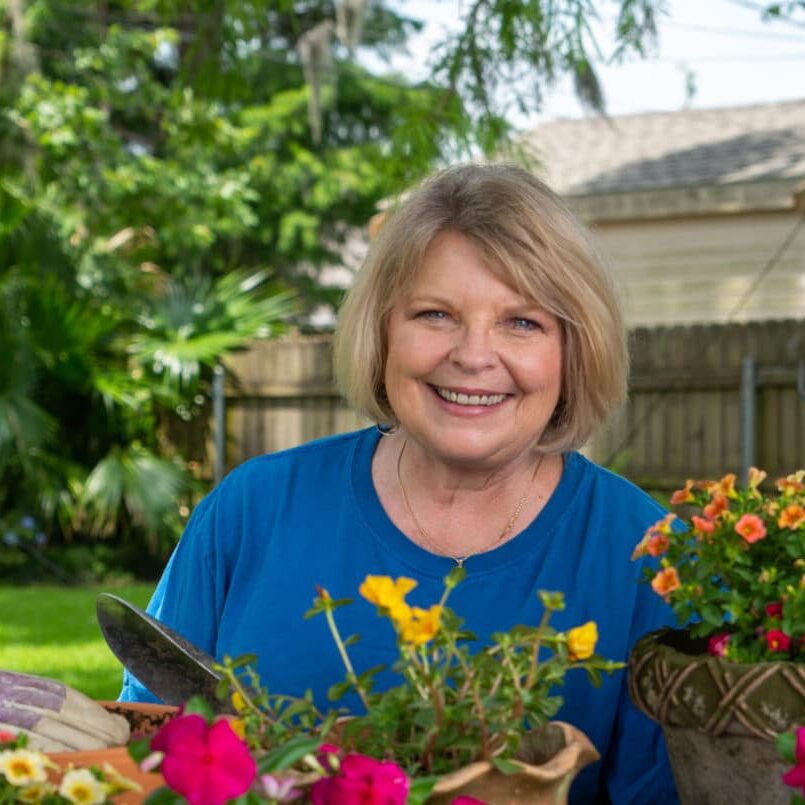 photo-1 A woman in a blue shirt standing in front of a pot of flowers.